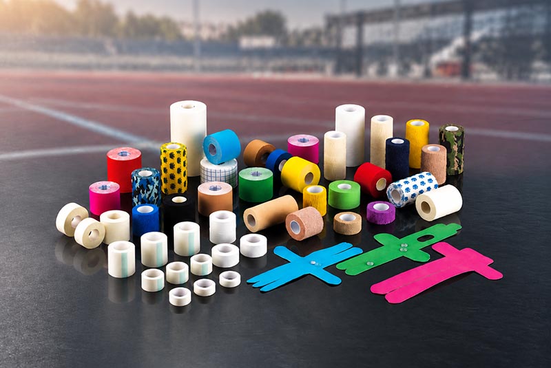 physiotherapy tape manufacturer displaying a range of colorful kinesiology tapes, rigid sports tapes, cohesive bandages and pre-cut athletic tapes against a blurred sports field background